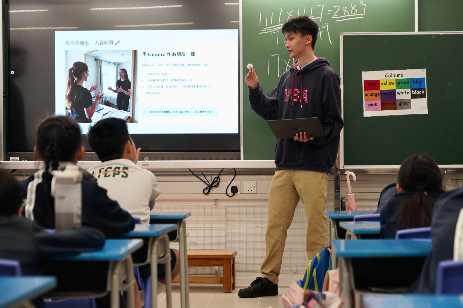 A tutor, laptop in hand, shows pupils how to explain a maths idea aloud to an AI to check they have understood it.