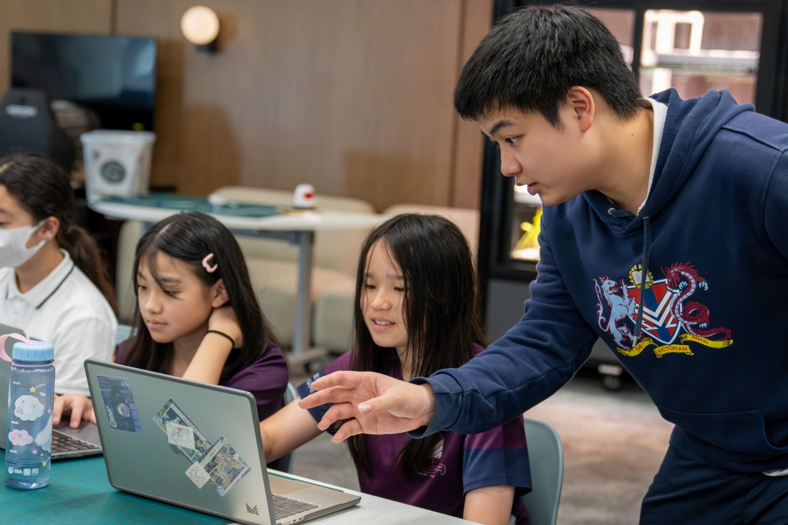 A tutor leans in to help two students working together at a laptop.
